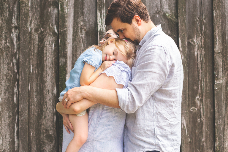 Family of 3 cuddling in front of barn wall | St. Louis Family Photos